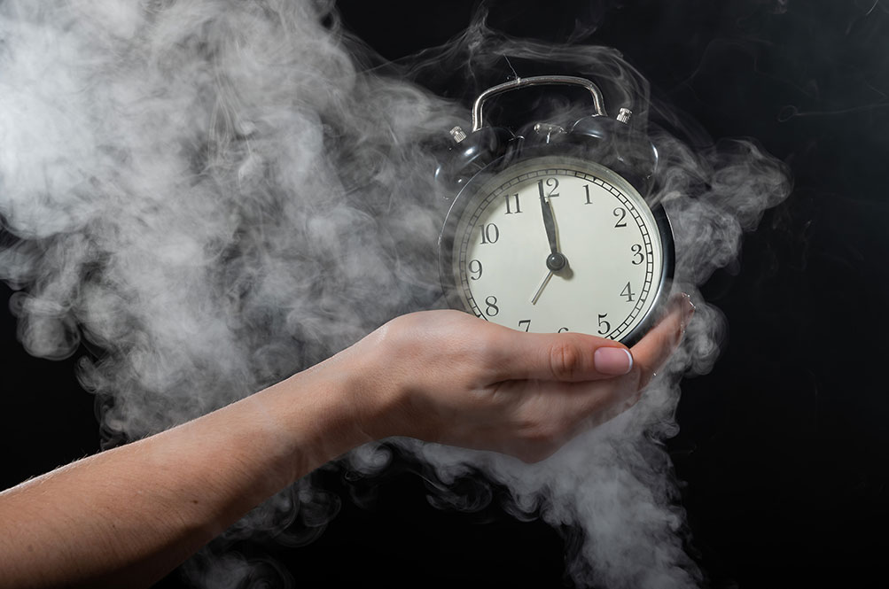 Close-up of a female hand holding a clock on a black background in smoke. Alarm clock at midnight in a mystical fog.